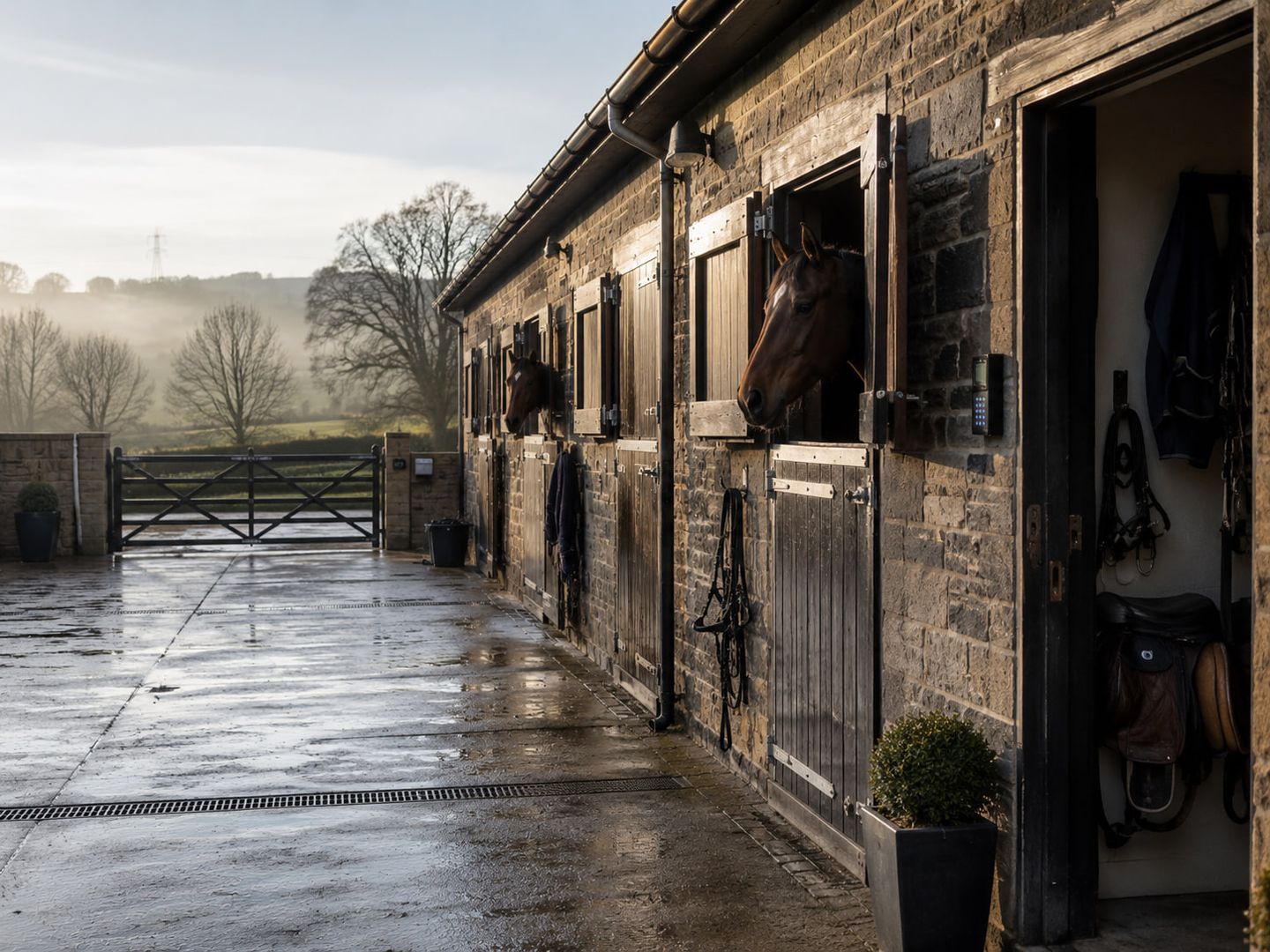 A stone stable block on a misty winter morning, two horses looking out over their doors onto a wet yard, with a five-bar gate and hills beyond.