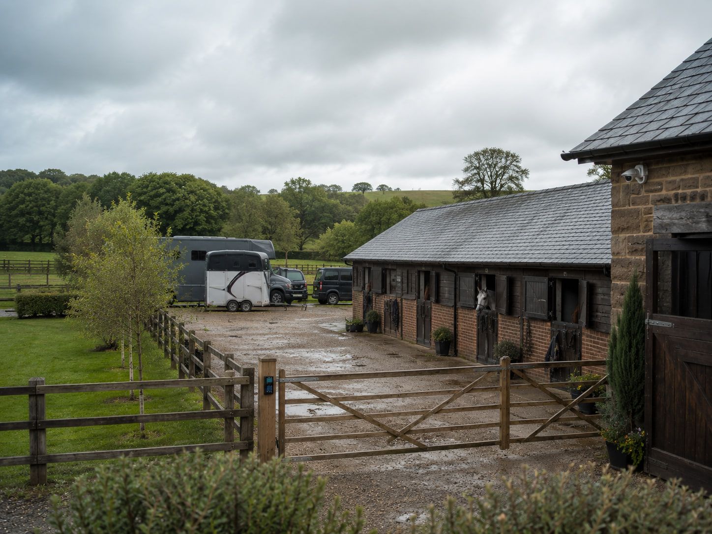 A stable yard under low cloud, a horsebox and trailer parked by the buildings and a timber five-bar gate closed across the entrance.