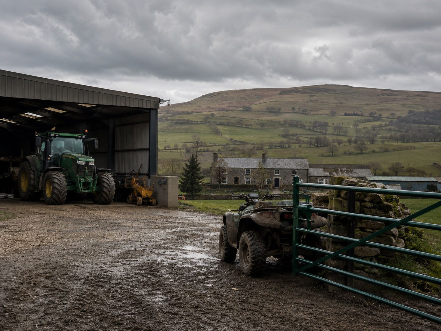 A working farmyard on a grey day, a tractor under an open machinery shed and a quad bike parked by a steel field gate, with hill grazing behind.