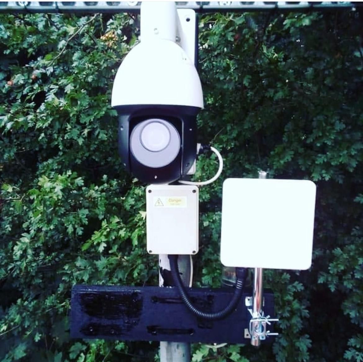 A pan-tilt-zoom camera and wireless antenna mounted together on a pole beside a hedgerow.