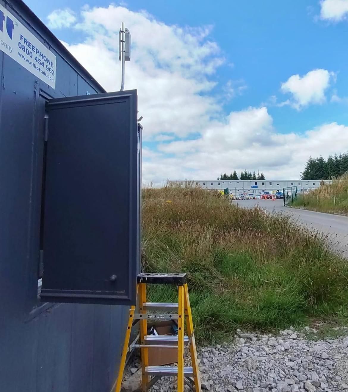A security cabinet and pole-mounted wireless antenna at an isolated site, with rough grazing and forestry behind.
